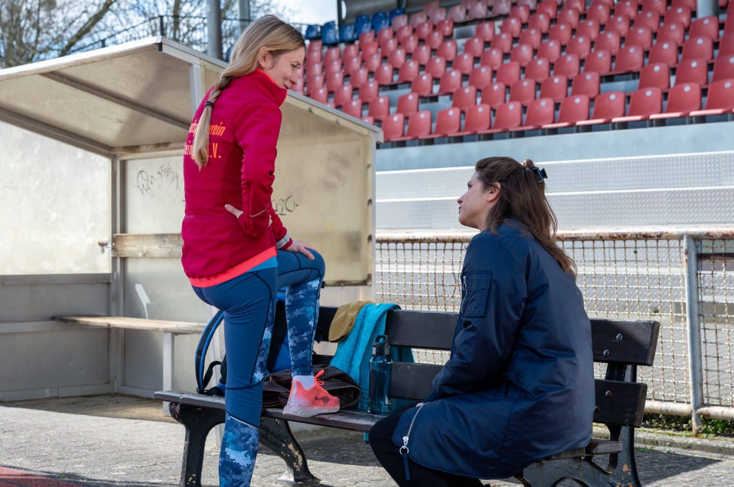 "Der Staatsanwalt - Verfeindet": Julia Schröder (Anika Baumann) sitzt am Rande eines Sportplatzes auf einer Bank und blickt zu Hannah Kurat (Marie Fischer), die in Sportkleidung ihr linkes Bein locker auf die Bank abstellt. Auf der Bank liegen ein Handtuch, eine Trinkflasche, eine Jacke und ein Rucksack. Hinter den beiden Frauen ist eine Zuschauertribüne mit roten Sitzen.
