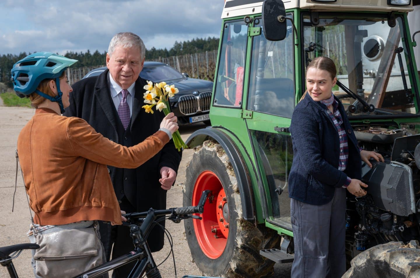 "Der Staatsanwalt - Verfeindet": Bernd Reuther (Rainer Hunold) und Luzie Monning (Melissa Maria Becker) stehen neben einem grünen Traktor und sehen beide zu Nora Monning (Paula Kroh), die einen Blumenstrauß mit ihrer rechten Hand hochhält. Mit ihrer linken Hand hält sie ein Fahrrad fest, auf dem Kopf trägt sie einen blauen Fahrradhelm. Einige Schritte hinter Reuther steht ein schwarzes Auto. Luzie Monning hat ihre Hände beide am Traktor, sie scheint etwas zu reparieren.
