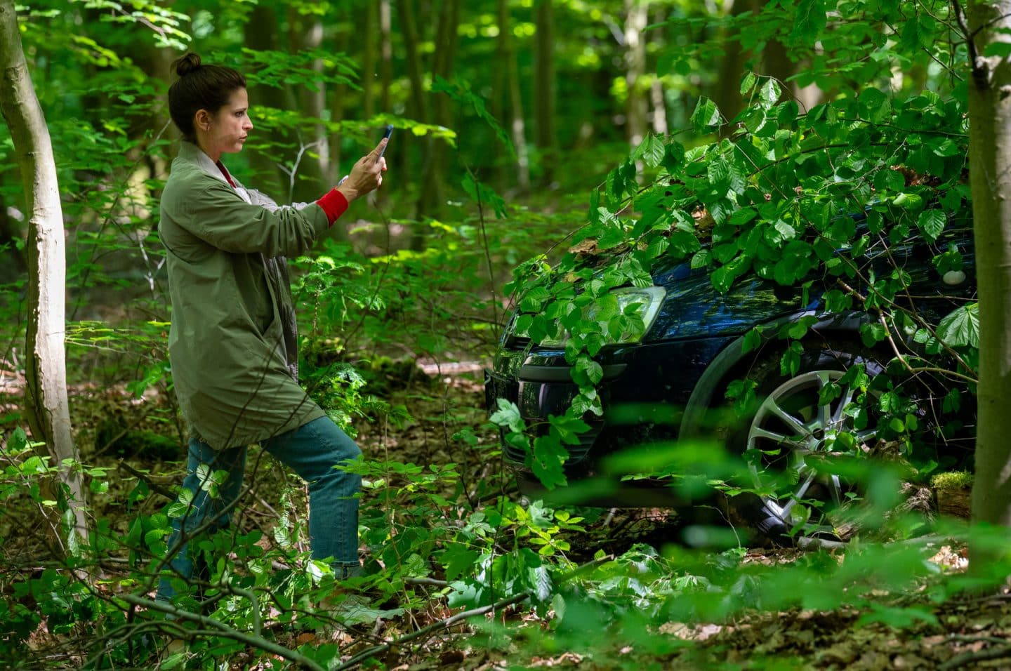 "Der Staatsanwalt - Tödliche Nähe": Julia Schröder (Anika Baumann) befindet sich im Wald. Sie hält mit ausgestreckten Armen ein Smartphone in den Händen. Vor ihr ist der vordere Teil eines Autos im Gebüsch zu erkennen.
