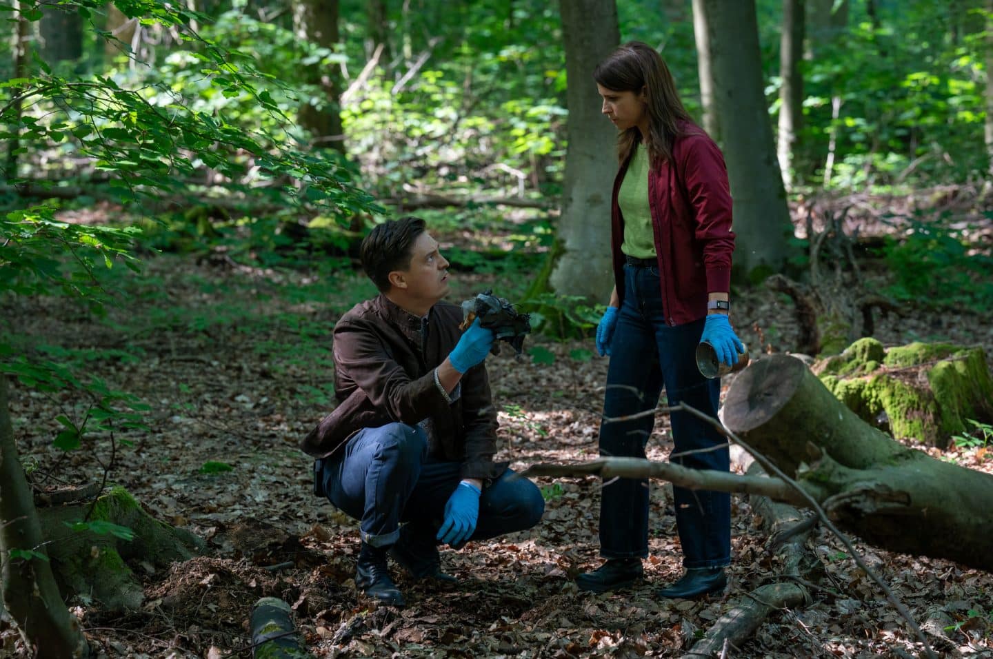 "Der Staatsanwalt - Zu allem entschlossen": Alexander Witte (Jannik Mioducki) kniet am Waldboden und hält einige Objekte in seiner rechten Hand. Julia Schröder (Anika Baumann) geht auf ihn zu und hält auch einen Gegenstand in ihrer linken Hand. Beide tragen blaue Handschuhe.
