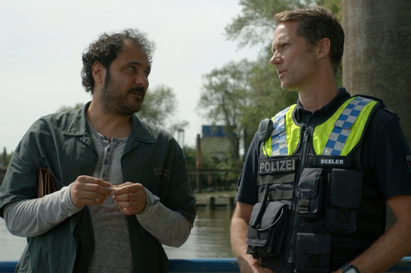 "Notruf Hafenkante - Kein Platz auf dieser Welt". Erhan Akin (Ali Berber) und der Polizist Mattes Seeler (Matthias Schloo) stehen nebeneinander an einer Brüstung am Fluss. Beide schauen sich an. Erkan ist im Begriff eine Zigarette zu drehen.