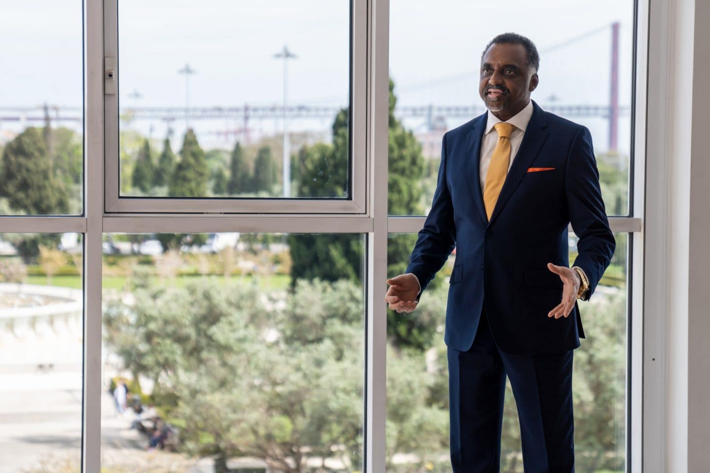 "Flucht aus Lissabon": Der kitwanesische Botschafter Idris Lumbada (Miguel Hurst) steht vor einer Fensterfront, die auf Bäume und Baukräne blicken lässt. Seine Gesten mit der Hand und sein Gesichtsausdruck wirken angespannt.
