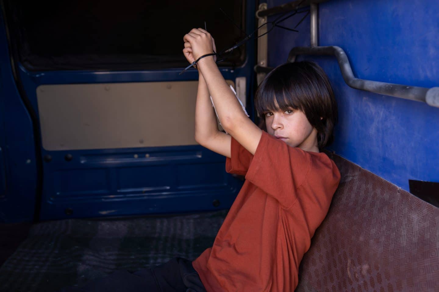 "Flucht aus Lissabon": Der junge Noa (Santiago André) sitzt im Inneren eines blauen Lieferwagens. Seine Hände sind mit Kabelbindern an die Autowand gebunden. Auf seinem rechten Unterarm klebt ein großes, weißes Pflaster. Sein entschlossener Blick geht in Richtung der Türen.
