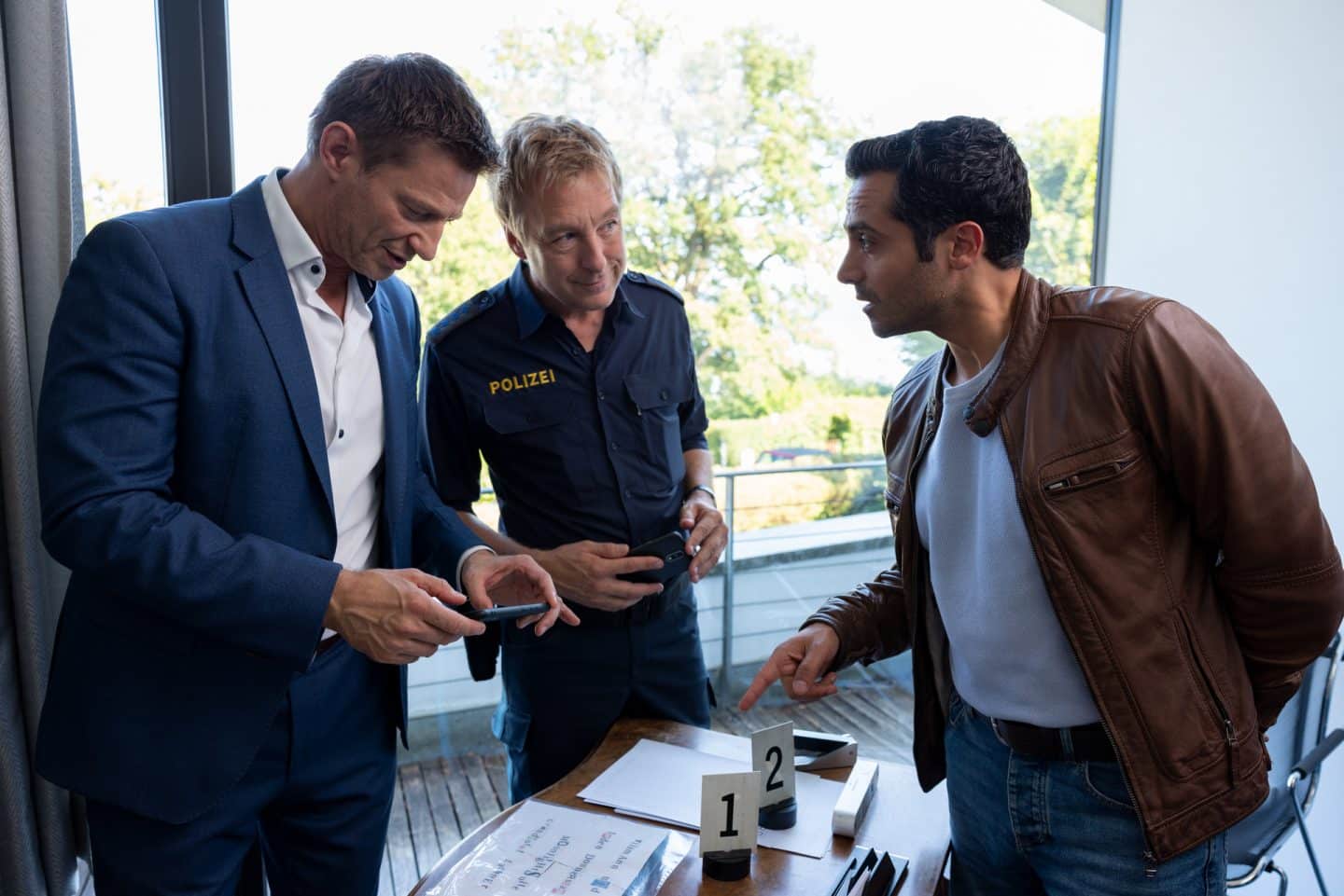 "Die Rosenheim-Cops - eine unglaubliche Affäre": Kommissar Hansen (Igor Jeftić), Polizeihauptmeister Mohr (Max Müller) und Kommissar Kaya (Baran Hêvî) stehen um einen Schreibtisch herum und unterhalten sich. Hansen fotografiert mit dem Smartphone ein Schreiben, das auf der Tischplatte liegt.
