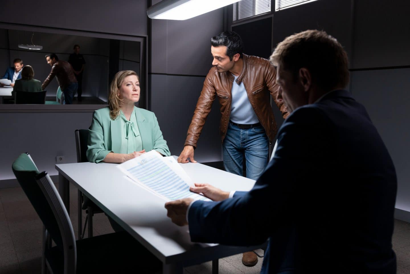 "Die Rosenheim-Cops - Eine hochintelligente Gesellschaft": Dr. Almuth Fehrenbacher (Ines Honsel) sitzt im Vernehmungsraum und sieht Kommissar Hansen (Igor Jeftić) an, der ihr gegenübersitzt und ein Dokument in den Händen hält, auf das er schaut. Neben Frau Dr. Fehrenbacher steht Kommissar Kaya (Baran Hêvî), der die Hand auf die Tischplatte gestützt hat, während er sie ansieht.