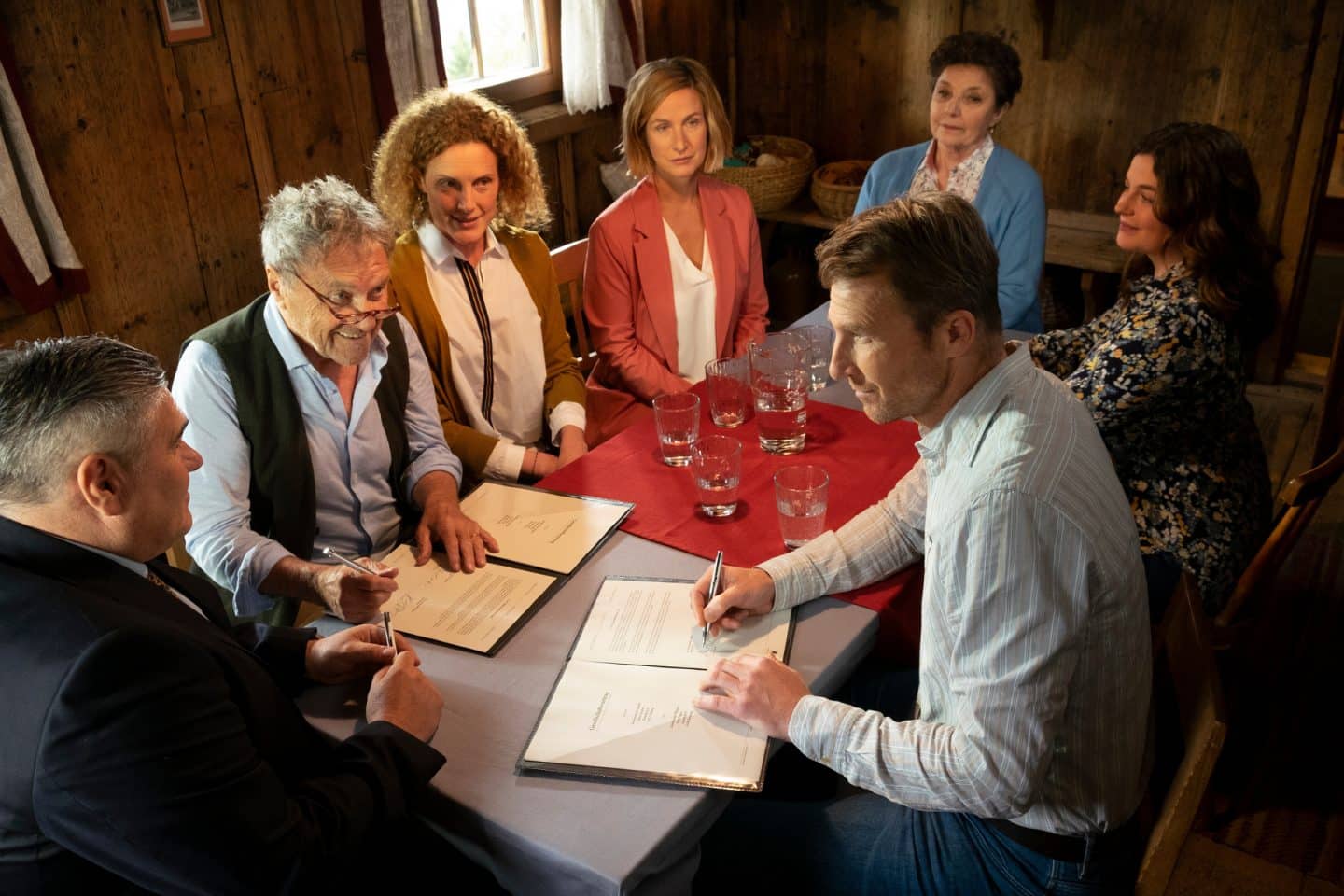 "Der Bergdoktor: Wechselwirkungen": Auf dem Gruberhof: An einem großen Tisch sitzen Hans (Heiko Ruprecht), Lilli (Ronja Forcher) und Lisbeth Gruber (Monika Baumgartner) sowie Caro (Barbara Lanz) und Rolf Pflüger (Wolfram Berger), außerdem Julia Andermatt (Nathalie Schott) und der Notar (Gerhard Salchner). Hans Gruber und Rolf Pflüger unterzeichnen vor ihnen liegende Papiere. Alle wirken zufrieden.