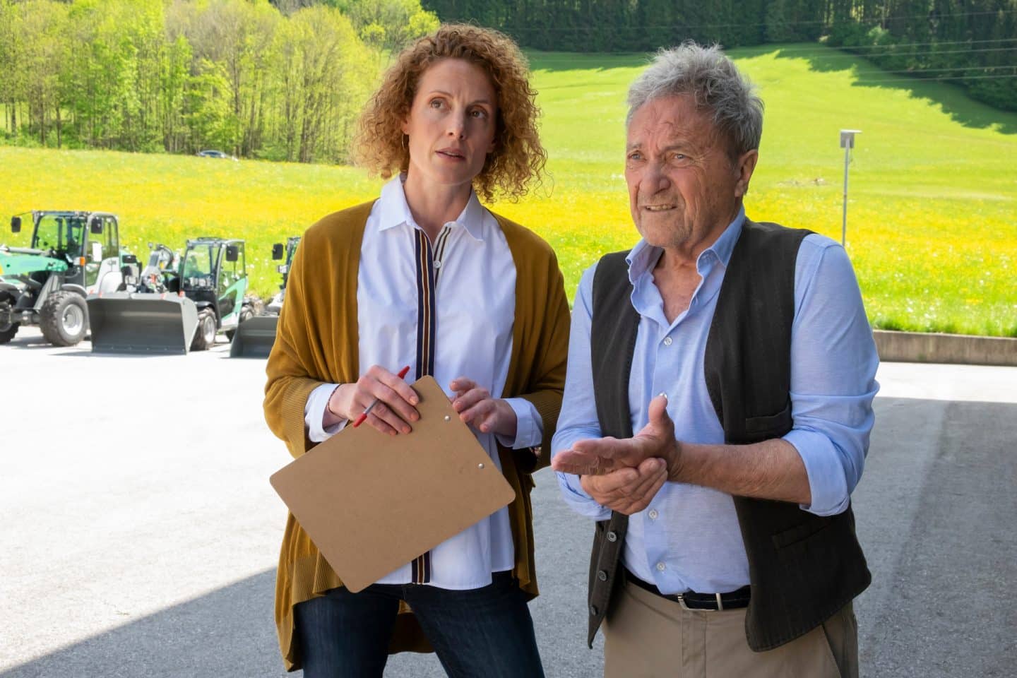 "Der Bergdoktor: Wechselwirkungen": Auf dem Hof des Landhandels: Caro Pflüger (Barbara Lanz) steht neben ihrem Vater Rolf Pflüger (Wolfram Berger). Sie hält ein Klemmbrett in den Händen und blickt fragend in die Ferne. Ihr Vater reibt sich lächelnd die Hände.