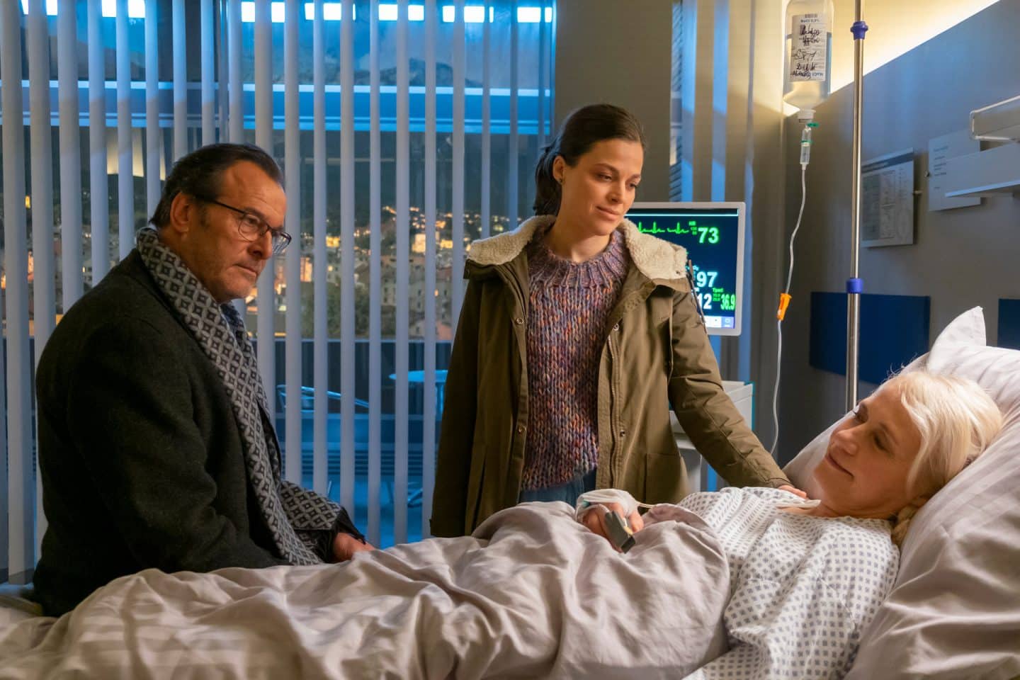 "Der Bergdoktor: Herzschmerzen": In der Klinik: Helen Aichinger (Alexandra von Schwerin) liegt im Krankenbett und ist an einer Infusion angeschlossen. Gerry Aichinger (Joachim Raaf) sitzt auf der Bettkante, Christina Aichinger (Giulia Goldammer) steht neben dem Bett. Alle wirken erleichtert.