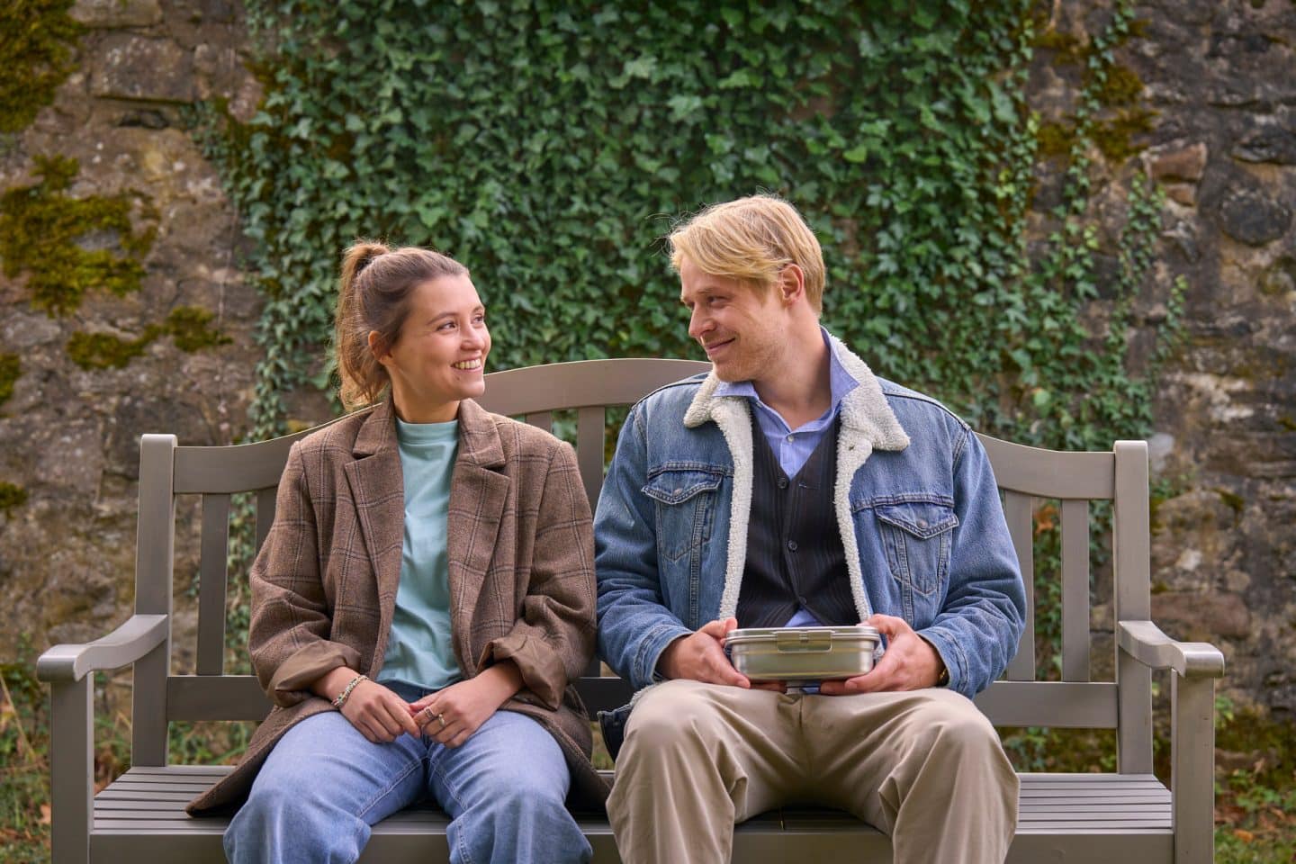 "Zimmer im Grünen - Herzenswege": Lisa (Janina Fautz) und Gabriel (Karl Schaper) sitzen auf einer Gartenbank vor einer mit Efeu bewachsenen Bruchsteinmauer. Sie lächeln sich an, Gabriel hält eine Brotbox in seinen Händen.