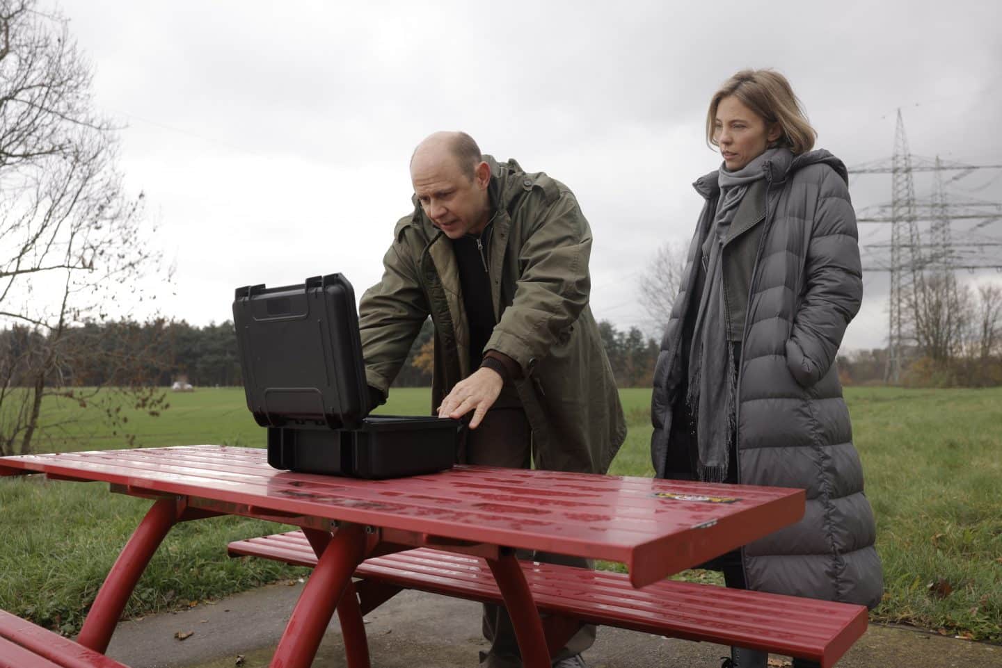"Yvonne und der Tod": Tim Koller (Moritz Führmann) steht vor einem Tisch auf einem Autobahnrastplatz. Er hat einen Koffer vor sich abgestellt und hantiert damit. Neben ihm betrachtet Conny Roth (Sina Bianca Hentschel) den Inhalt des Koffers.
