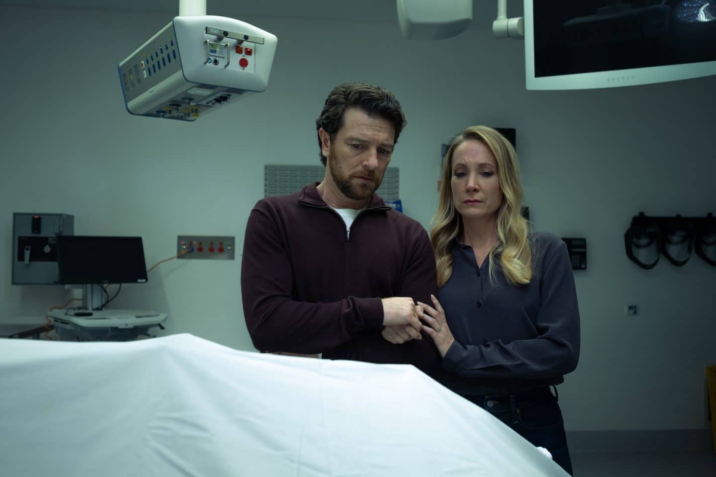 "North Shore – Tod in Sydney - Drummond Down Under": Abigail Crawford (Joanne Froggatt) und ihr Mann Simon Chalcott (Dan Spielman) stehen nebeneinander in einem Raum in der Gerichtsmedizin. Vor ihnen auf dem Tisch liegt eine Leiche unter einem weißen Tuch. Abigail hält Simons Hand.