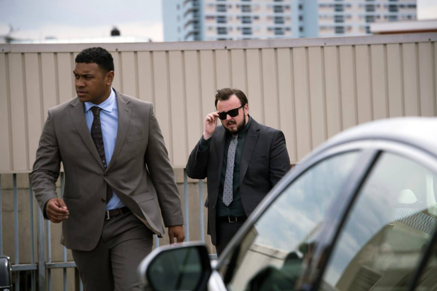 "North Shore – Tod in Sydney - Stanleygate": DC Paea Leniu (Chris Alosio) geht an einem geparkten Auto vorbei. DS Max Drummond (John Bradley) folgt ihm. Max fasst mit der rechten Hand an einen Bügel seiner Sonnenbrille.