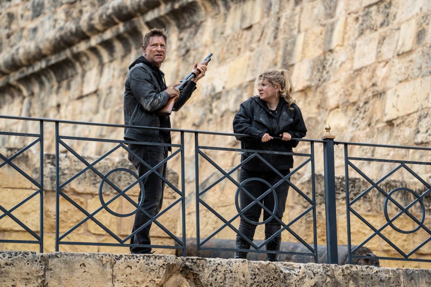 "Geheimsache Malta": Shaw (Aaron Eckhart) und MI6-Analystin Kacey (Abigail Breslin) stehen nebeneinander an einem Geländer vor den Mauern eines alten Gebäudes. Shaw hält ein Gewehr in den Händen und sieht in die Ferne. Kacey umklammert mit ihren Händen das Geländer und sieht ihn an.