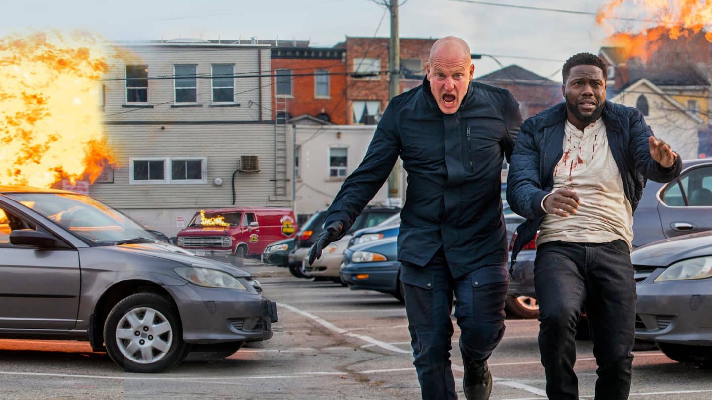 "The Man From Toronto": Der Mann aus Toronto (Woody Harrelson) und Teddy (Kevin Hart) rennen über einen Parkplatz, während im Hintergrund mehrere Gebäude explodieren.
