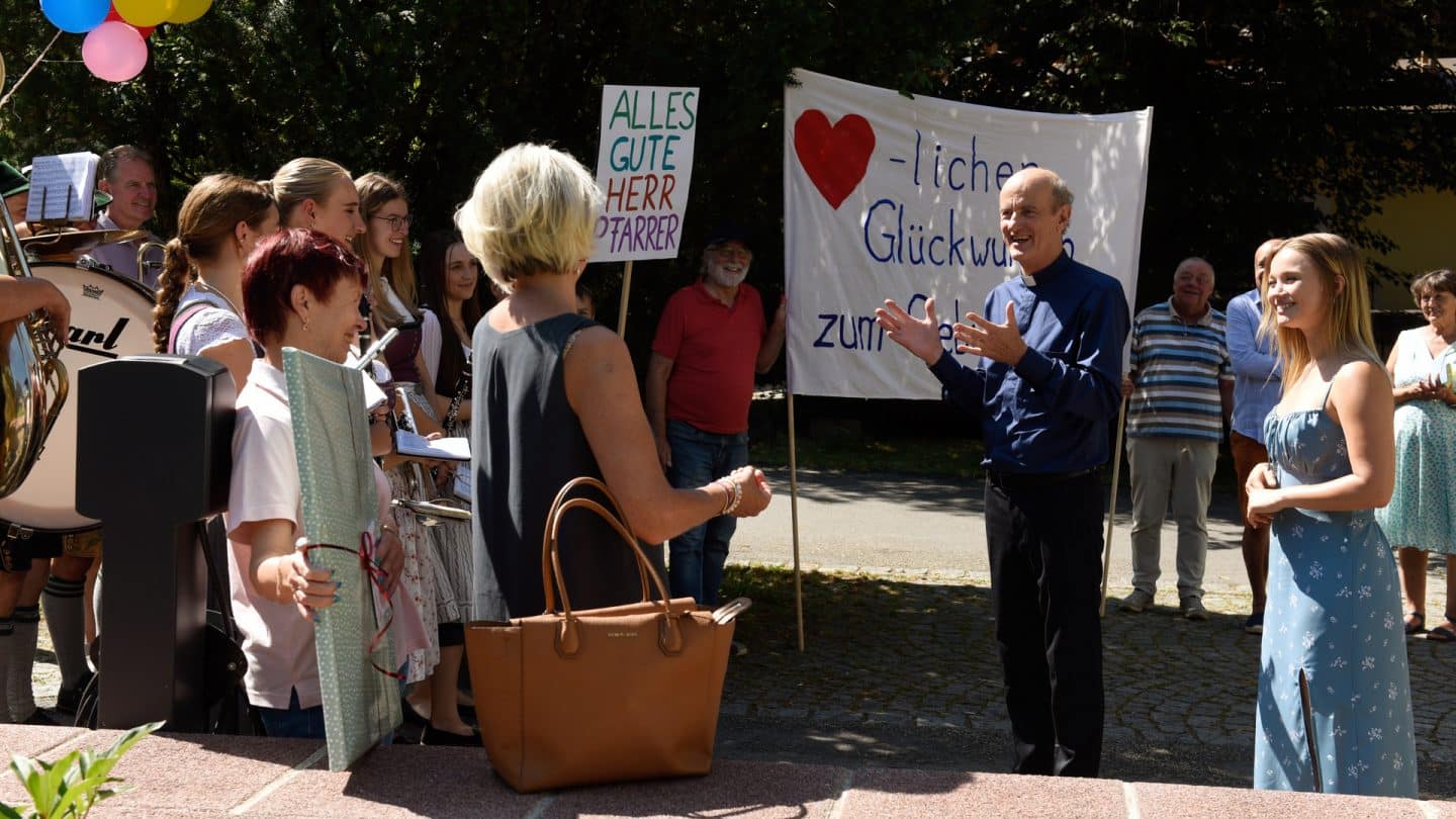 "Frühling - Babyalarm": Im Freien: Pfarrer Sonnleitner (Johannes Herrschmann) und Lilly Engel (Julia Willecke) sind umringt von einer kleinen Menschenmenge. Die Bewohner Frühlings halten Schilder in die Höhe, auf denen "Alles Gute Herr Pfarrer" und "Herzlicher Glückwunsch" geschrieben steht.
