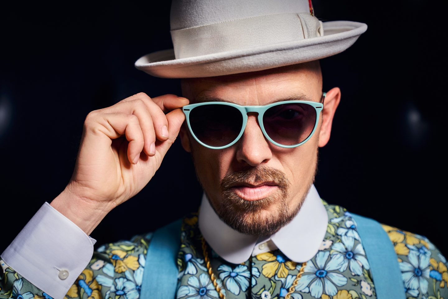 Jan Delay mit weißem Hut und mintgrüner Sonnenbrille. Der Musiker trägt ein buntes, florales Hemd mit blauen Hosenträgern und schaut ernst in die Kamera