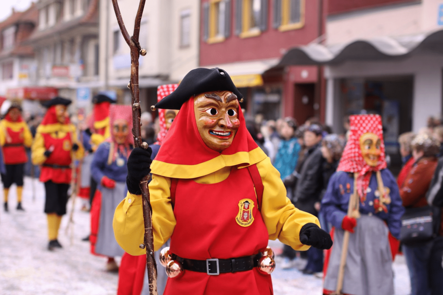Person in einem traditionellen Fasnetskostüm mit roter und gelber Tracht, schwarzer Dreispitzmütze und geschnitzter Holzmaske mit freundlichem Ausdruck. In der Hand ein geschmückter Stock, am Gürtel hängen Glocken. Im Hintergrund weitere verkleidete Personen in ähnlichen Kostümen sowie Zuschauer entlang der Straße.







