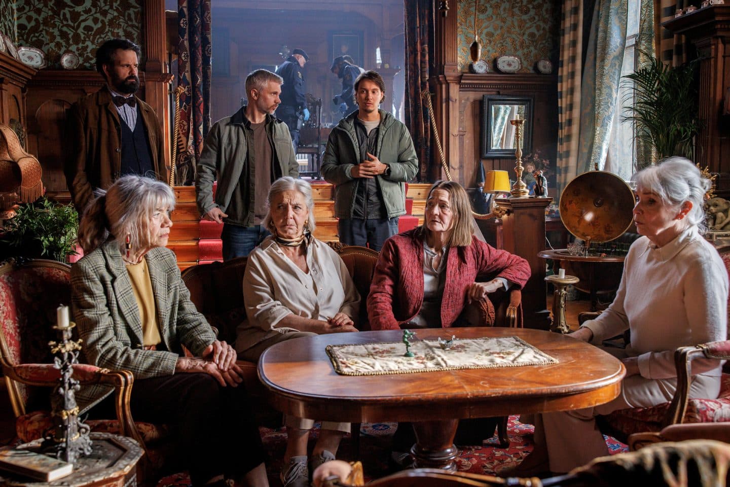 "Mord im Mittsommer - Birgitta": Kerstin (Iwa Boman), Monica (Louise Edlind Friberg), Ursula (Ann Petrén) und Anita (Marika Lindström) sitzen in einem antik eingerichteten Wohnraum um einen Holztisch herum. Im Hintergrund stehen Tony (Eric Stern), Alexander (Nicolai Cleve Broch) und Welpe (Julius Fleischanderl).
