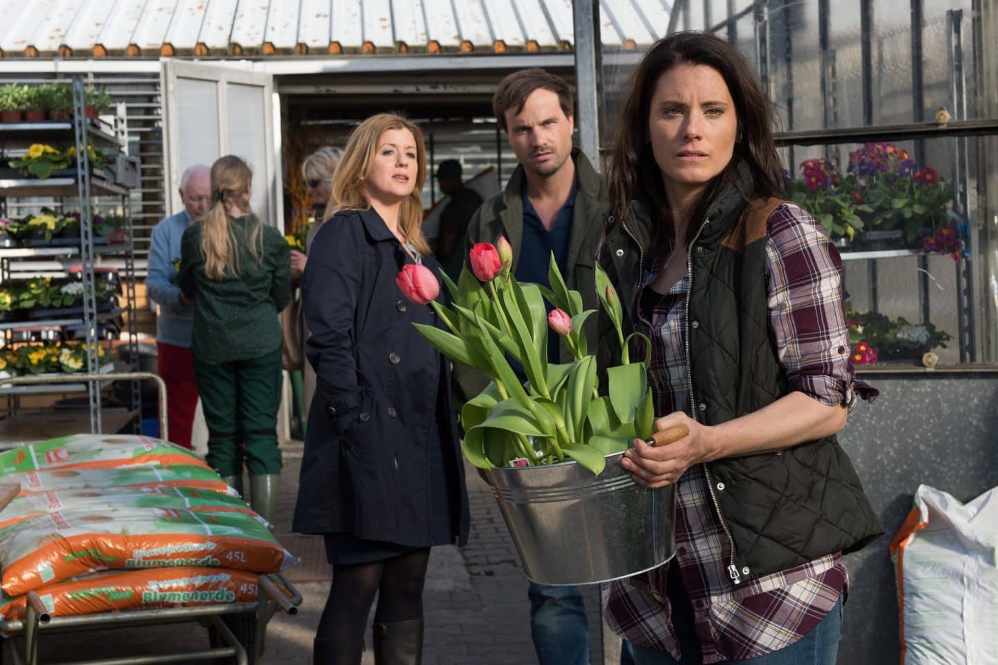 "Der Staatsanwalt – Die Macht der Sterne": Kerstin Klar (Fiona Coors) und Christian Schubert (Simon Eckert) stehen nebeneinander in einer Gärtnerei. Sie schauen zu Antonia Rumpf (Elisabeth Baulitz), die von ihnen abgewandt steht und einen Topf mit Tulpen in Händen hält.