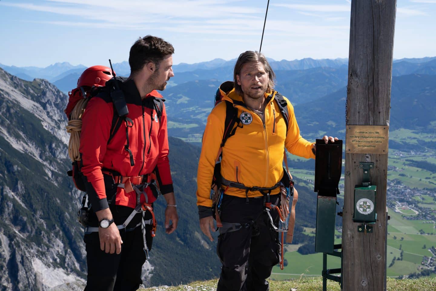"Die Bergretter: Im Gipfelbuch": Die Bergretter Markus (Sebastian Ströbel) und Simon (Ferdinand Seebacher) stehen am Gipfel eines Berges und wirken überrascht. Markus hält mit einer Hand eine Klappe fest, die normalerweise das Gipfelbuch sichert.
