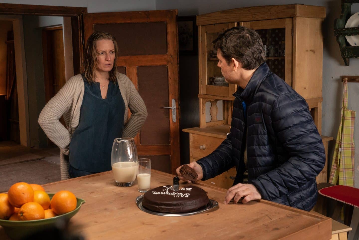 "Die Bergretter: Eiskalte Wahrheit (1)": Lorenz (Steffen Groth) sitzt an einem Tisch auf dem eine Karaffe mit Milch sowie ein Kuchen steht. Er hält dabei ein Stück Kuchen in seiner Hand und blickt in Emilies (Stefanie von Poser) Richtung. Diese steht daneben und blickt mit nassen Haaren und Händen in der Hüfte, verärgert in seine Richtung.