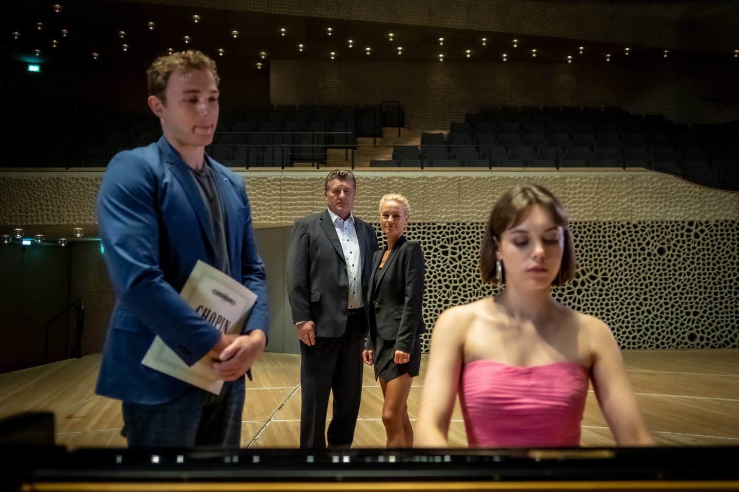 "Notruf Hafenkante - Die große Chance": Hannah Jacobi (Valerie Stoll) sitzt an einem Flügel auf der Bühne der Elbphilharmonie. Neben ihr steht Thilo Bahnsen (Moritz von Zeddelmann), der ihr beim Spielen zusieht. Im Hintergrund stehen der Polizist Hans Moor (Bruno F. Apitz) und seine Kollegin Franzi Jung (Rhea Harder-Vennewald), beide in Zivil, und beobachten Hannah.
