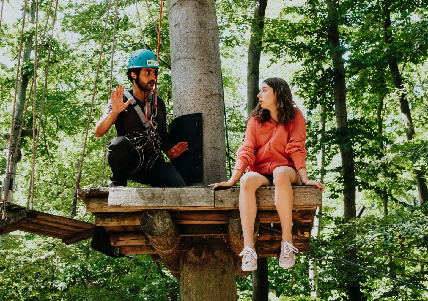 "SOKO Potsdam - Mutterliebe": Fine Nowak (Emilia Eidt) sitzt auf einer hohen Plattform aus Holz im Kletterwald. Sie sieht nach links zu David Grünbaum (Omar El-Saeidi), der neben ihr auf der Plattform hockt. Er ist mit einem Seil an einem Karabinerhaken gesichert, trägt einen blauen Helm und hat beschwichtigend seine Hände gehoben. Fine Nowak ist komplett ungesichert.