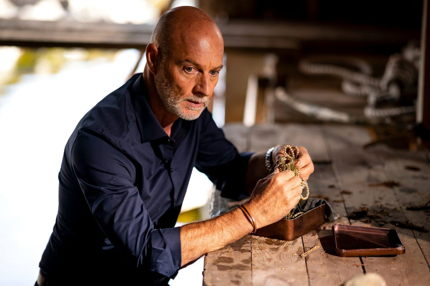 "Inga Lindström: Geliebter Feind": Viggo (Simon Licht) steht in einem Bootshaus aus Holz im Wasser. Auf einem Holzsteg vor ihm eine Metall-Schatulle mit altem Schmuck. Viggo schaut ergriffen und nachdenklich zur Seite.
