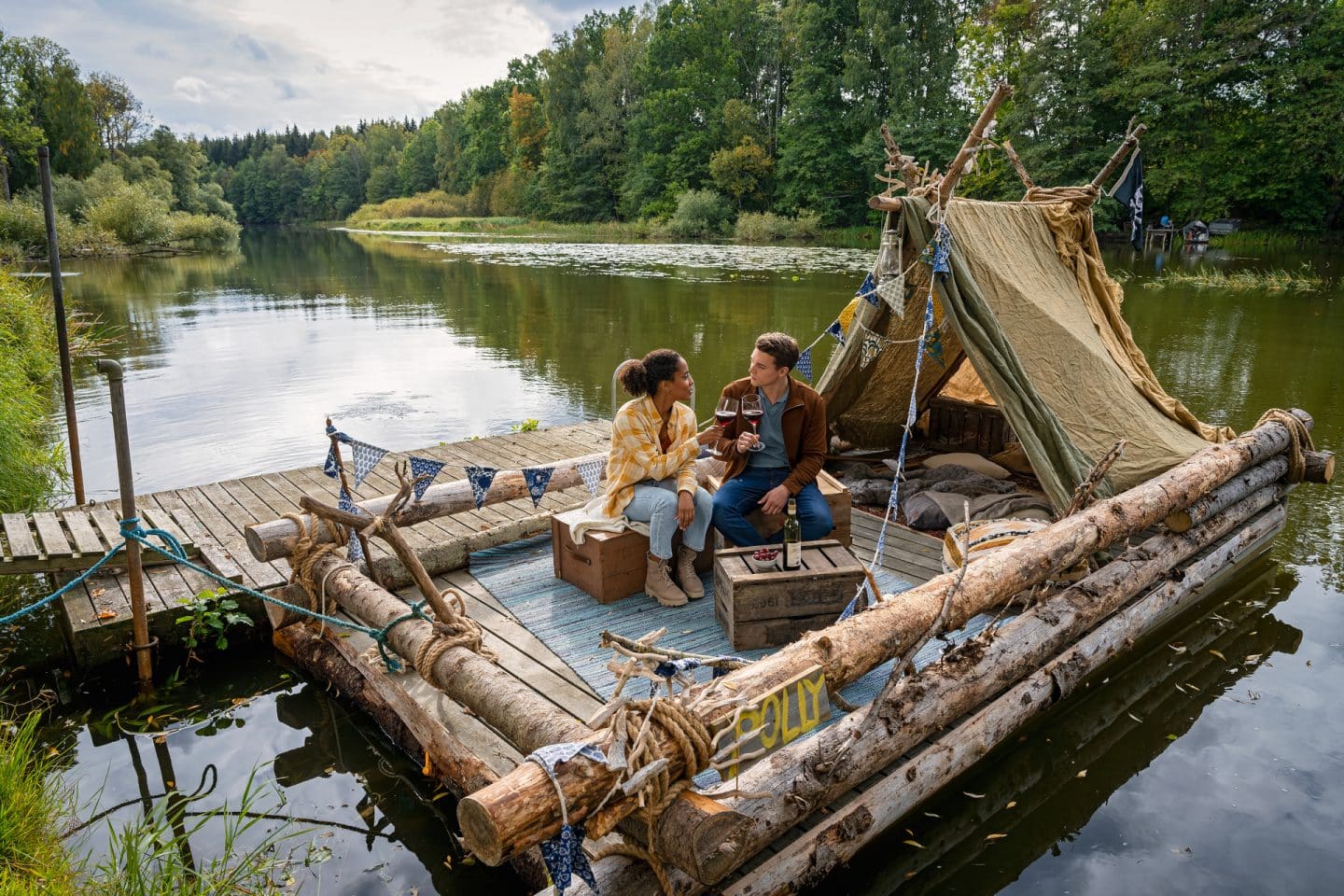 "Inga Lindström: Geliebter Feind": Elli (Pia Amofa-Antwi) und Lucas (Gerrit Klein) sitzen auf Kisten auf dem eben fertig gestellten Floß. Sie prosten sich mit Rotweingläsern zu und schauen sich an. Das Floß liegt an einem Holzsteg an einem idyllischen See im Grünen.
