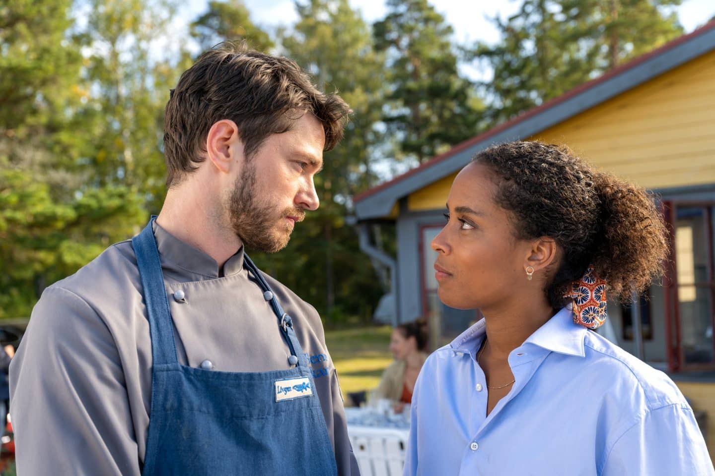 "Inga Lindström: Geliebter Feind": Elli (Pia Amofa-Antwi) und Benne (Josia Krug) stehen vor dem Gasthof der Lövgrens, der zwischen einem Wald und einem Seeufer steht. Beide blicken sich fragend an.
