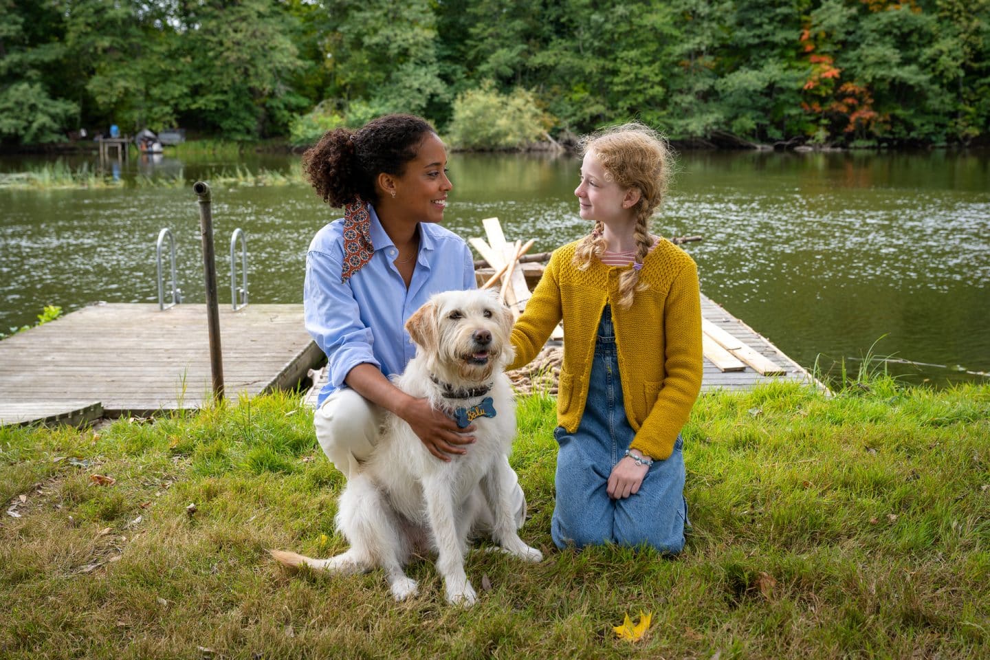 "Inga Lindström: Geliebter Feind": Elli (Pia Amofa-Antwi) und Ronja (Ida Wieland) knien auf einer Wiese, zwischen ihnen sitzt der Hund Balu. Beide schauen sich an und lächeln. Im Hintergrund ein See mit einem Holzsteg und einem unfertigen Floß.
