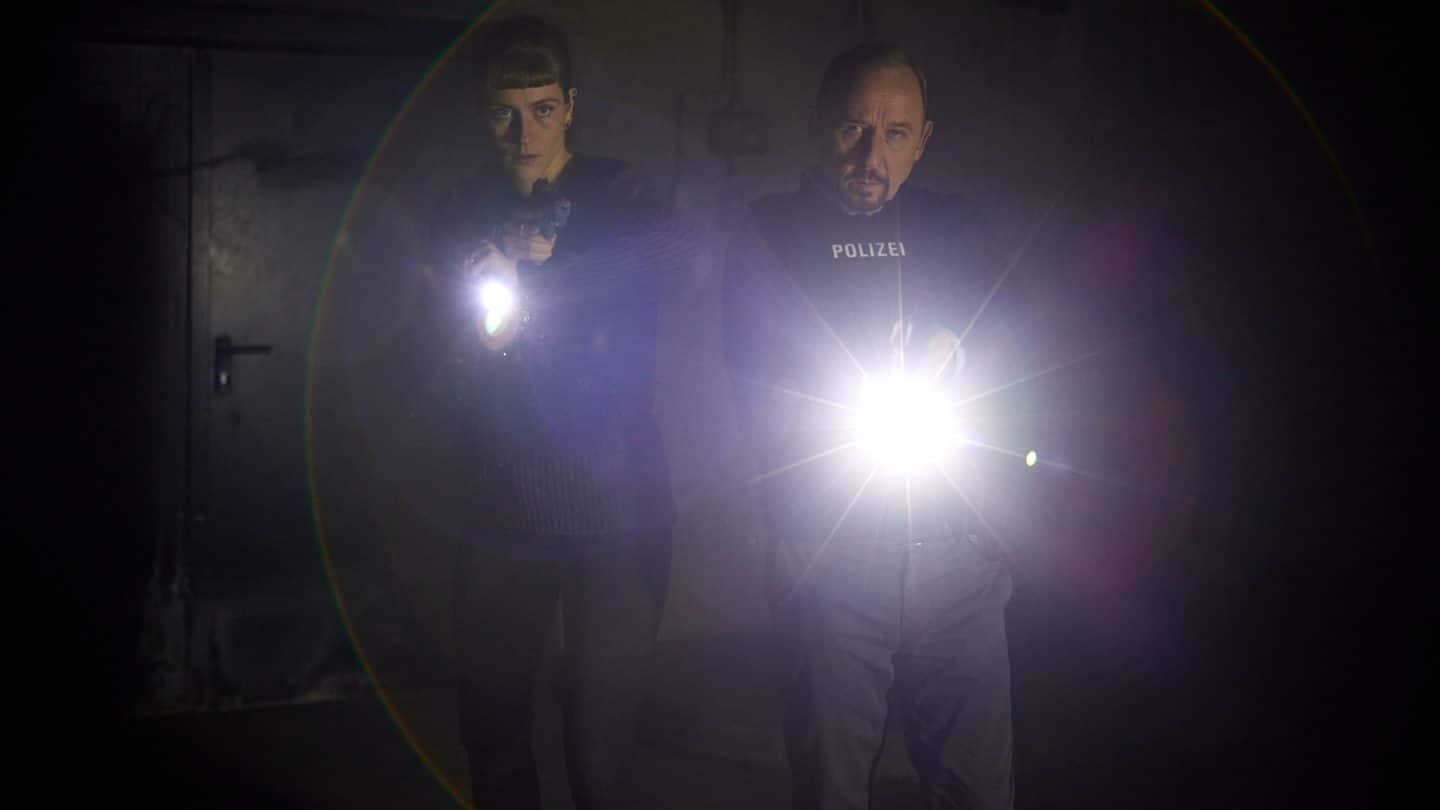 "Stralsund - Der letzte Sieg": Jule Zabek (Sophie Pfennigstorf) und Karl Hidde (Alexander Held) stehen mit gezogenen Waffen und schusssicheren Westen im Tunnelsystem. Sie leuchten mit ihren Taschenlampen nach vorne.