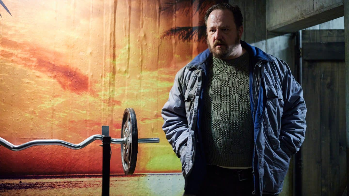 "Stralsund - Der letzte Sieg": Andy Block (Andreas Anke) steht vor einer Fototapete mit einem Sonnenuntergang. Neben ihm steht eine Stange zum Gewichtheben.
