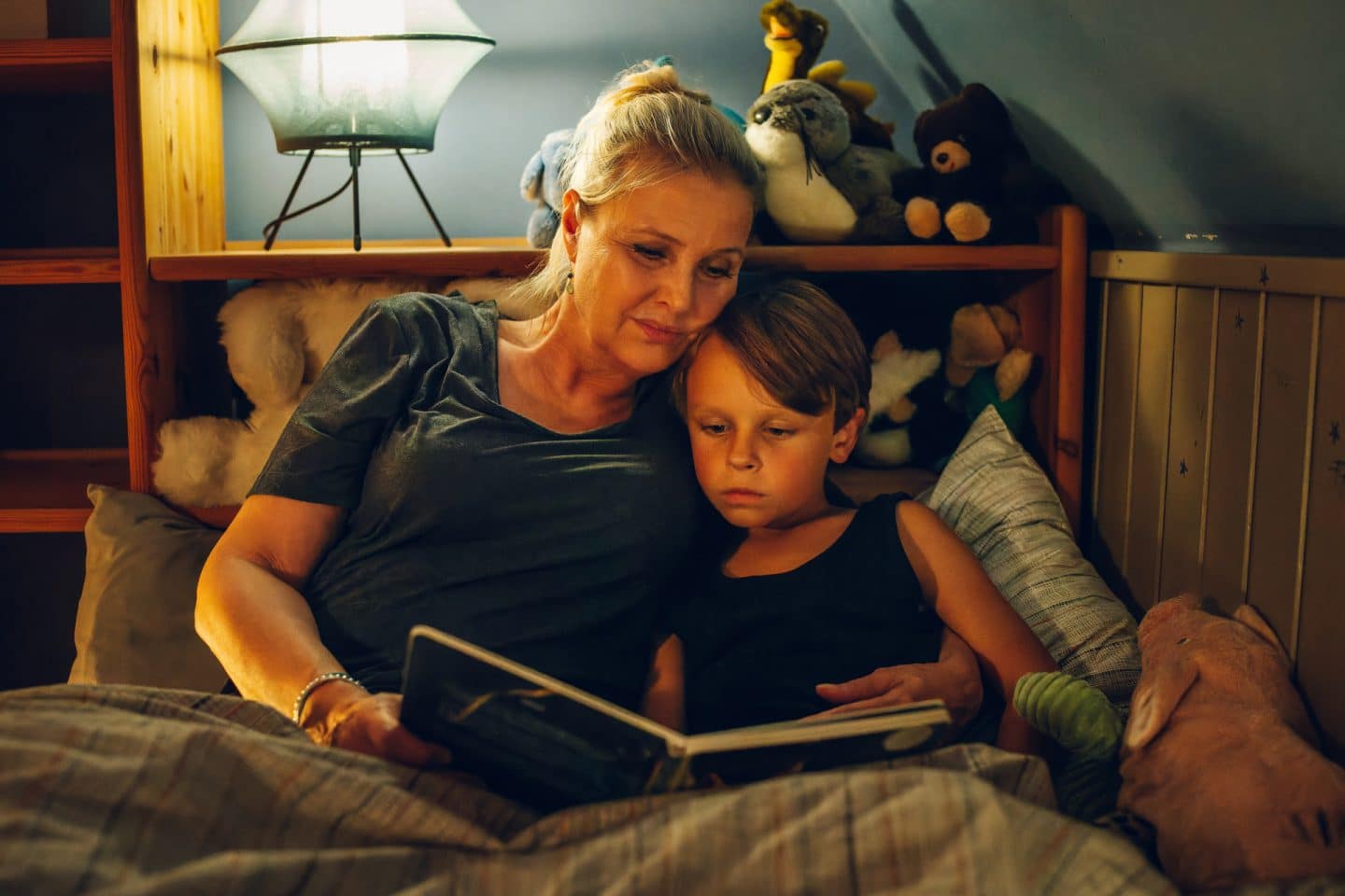 "Das gläserne Kind": Anne Schuchardt (Katharina Böhm) hat ihren Enkel Luke (Lennox Louis Steigerschmid) im Arm. Sie liegen gemeinsam im Bett und schauen in ein Buch. Im Hintergrund liegen viele Kuscheltiere neben einer Nachtischlampe.
