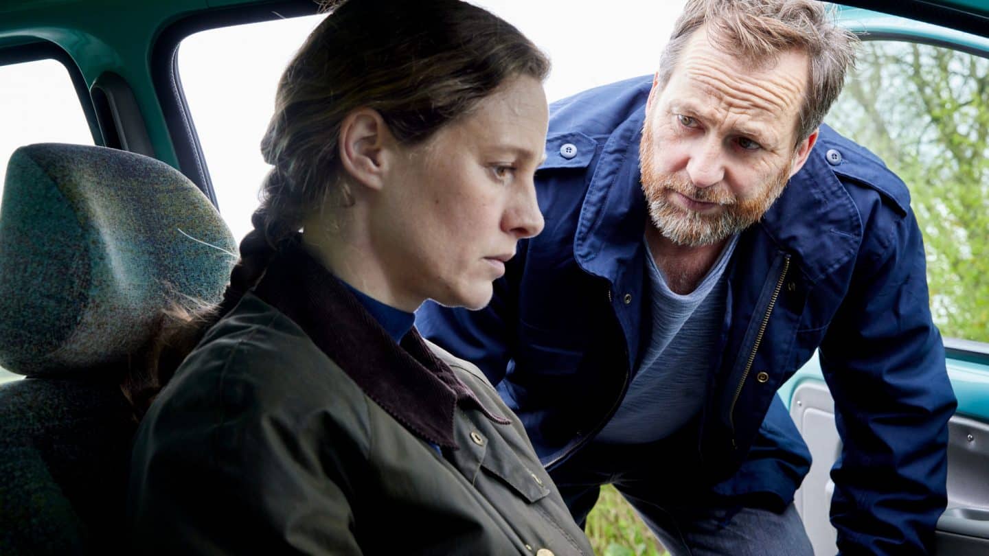 "Ostfriesenfluch": Frank Weller (Christian Erdmann) beugt sich mit besorgter Miene zu seiner Frau Ann Kathrin Klaasen (Picco von Groote) hinunter, die bedrückt auf dem Vordersitz des Autos sitzt und vor sich hinstarrt.
