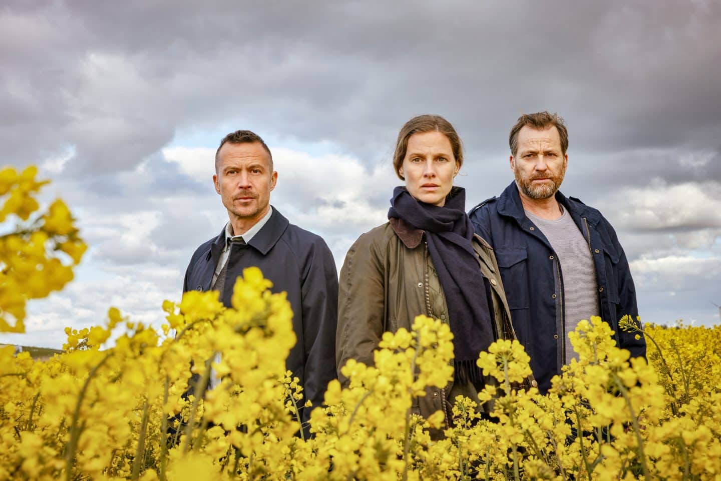 "Ostfriesenfluch": Rupert (Barnaby Metschurat,), Ann Kathrin Klaasen (Picco von Groote) und Frank Weller (Christian Erdmann) stehen mitten in einem gelben Rapsfeld etwas versetzt nebeneinander. Sie sind nur ab dem Oberkörper aufwärts zu sehen.
