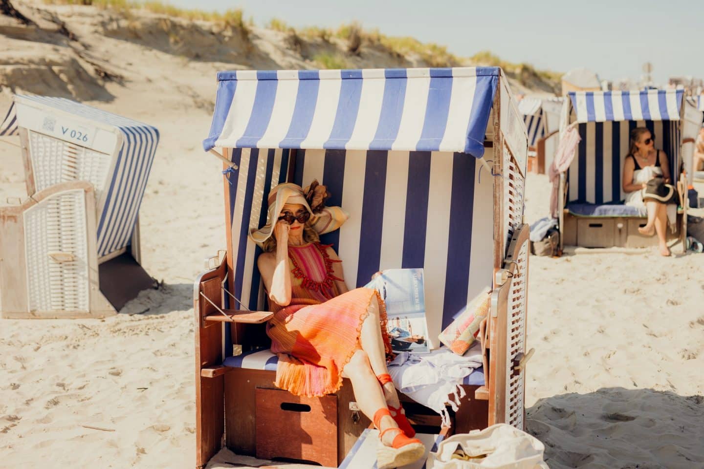 "Verhängnisvolle Leidenschaft Sylt": Am Stand: Hedi von Carlsberg (Leslie Malton) sitzt in einem Strandkorb. Sie hat die Beine übereinandergeschlagen und blickt, die Sonnenbrille leicht heruntergezogen, keck in die Kamera.
