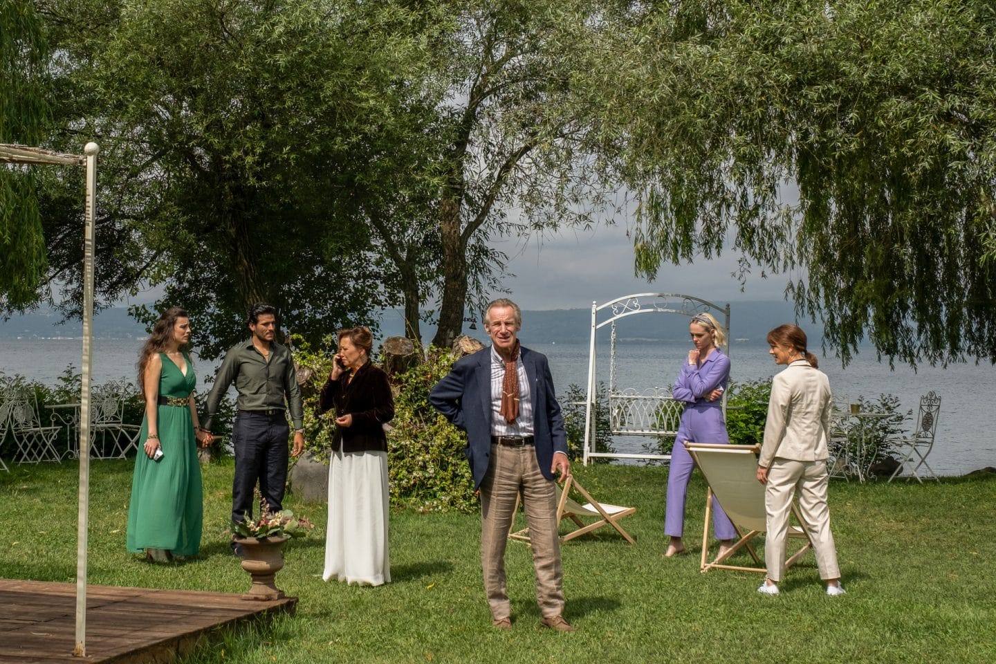 "Signora Volpe - Verstrickung": Luciana Garcia (Adriana Laespada), Ricardo Garcia (José Neira), Lee Hamilton (Fiona Marr), Michael Priest (Nicholas Farrell), Zoe Hamilton (Natalie Shinnick) und Sylvia Fox (Emilia Fox) alias "Kirsty" stehen nebeneinander in einem Garten. Im Hintergrund stehen große Bäume am Ufer eines Sees. Lee telefoniert.
