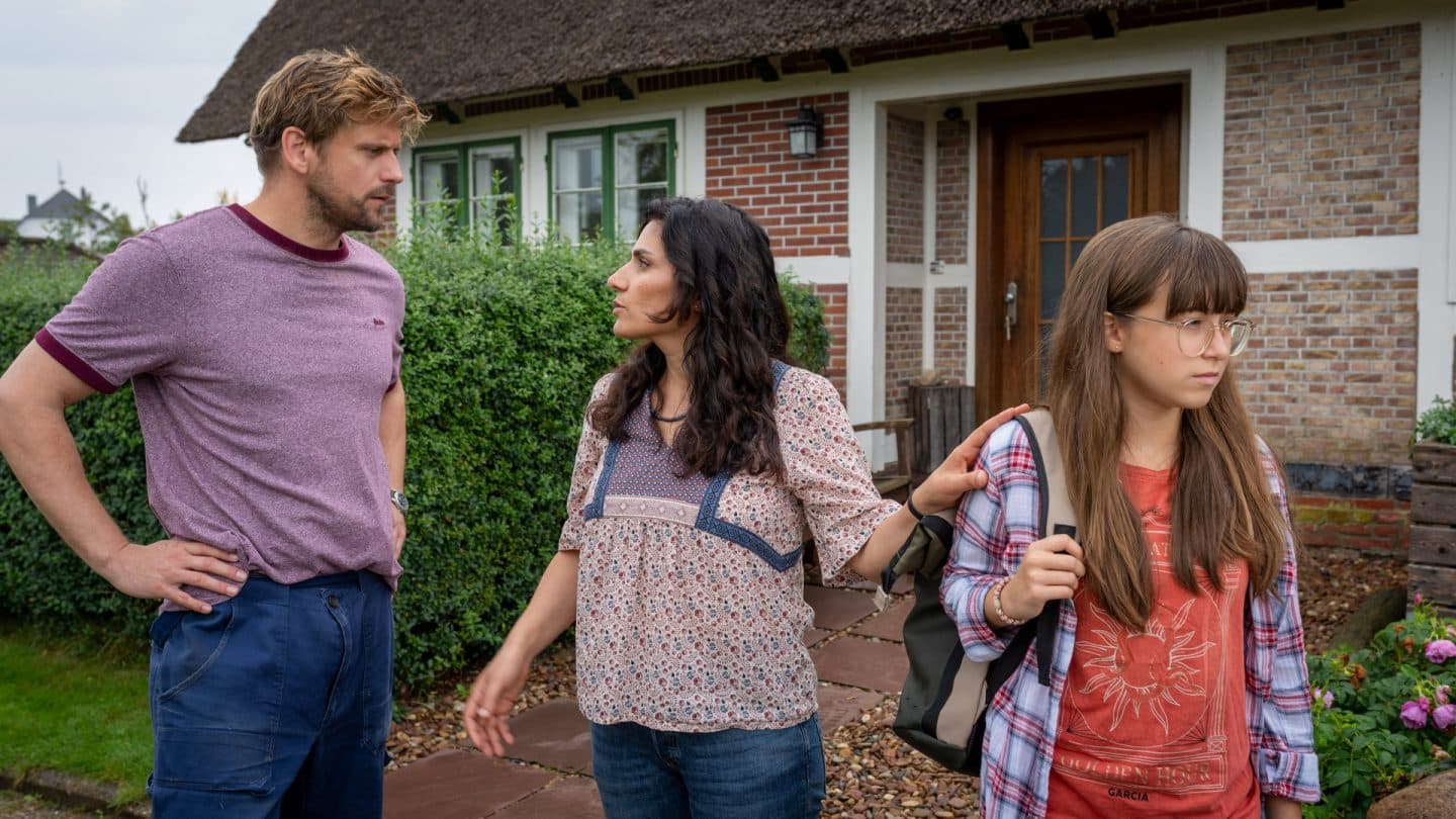 "Neuer Wind im Alten Land - Erntezeit": Vorm Haus steht Paul Harms (Steve Windolf) Elif Baskan (Halima Ilter) mit strengem Blick gegenüber. Von beiden abgewandt steht Laila (Elsa Krieger). Die Hand ihrer Mutter liegt auf ihrer Schulter.
