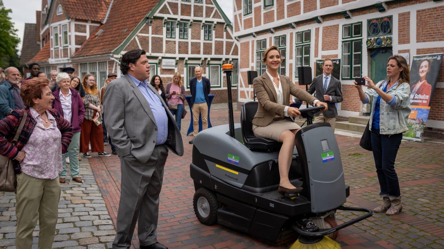 "Neuer Wind im Alten Land - Erntezeit": Edeltraud Fock (Elga Schütz) und Elmar Bröhan (Michael Rothmann) schauen zu wie Heide Schulze (Anne Roemeth) die neue Kehrmaschine vorführt. Beke Rieper (Felicitas Woll) hält unterdessen alles mit dem Handy fest. Im Hintergrund stehen Passanten (Komparsen) vor Fachwerkhäusern.
