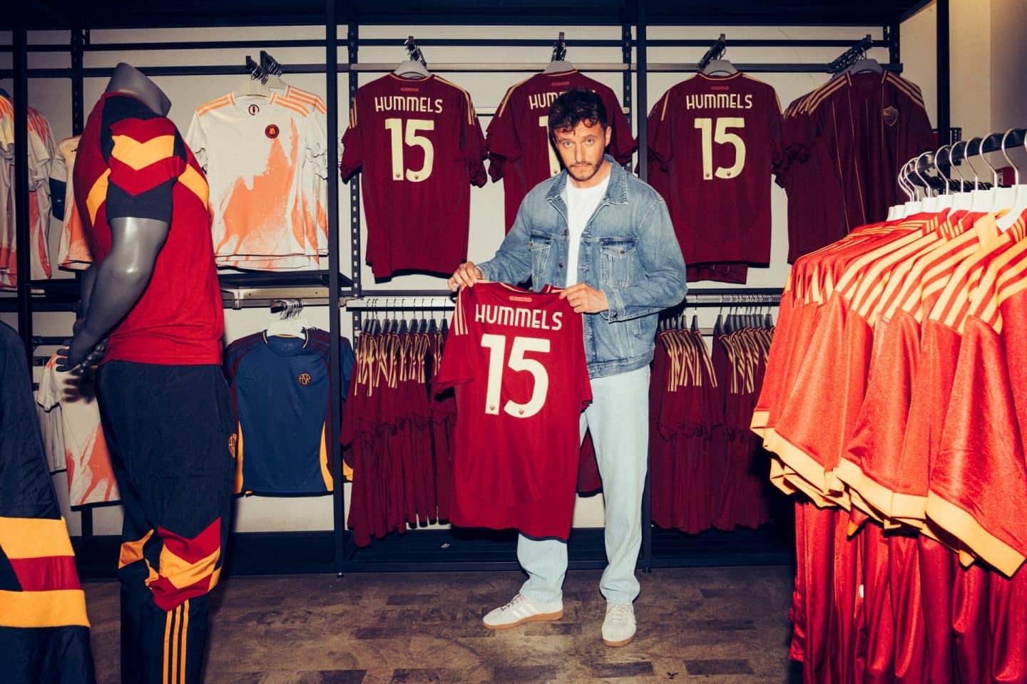 "Hummels - La Finale. Mit Tommi Schmitt": Tommi Schmitt steht in einem Trikot-Sportgeschäft. Es hängen mehrere Hummels Trikots im Aushang. Tommi Schmitt hält eines der Trikots mit der Nummer 15 und Hummels-Aufdruck in die Kamera.
