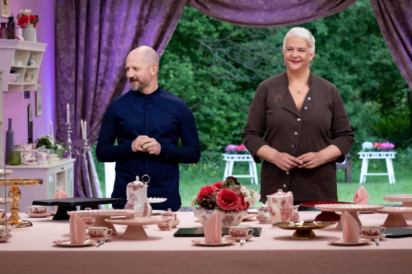 Christian Hümbs und Bettina Schliephake-Burchardt stehen als Jury von „Das große Backen“ hinter einem festlich gedeckten Tisch mit Tortenplatten und Porzellan.