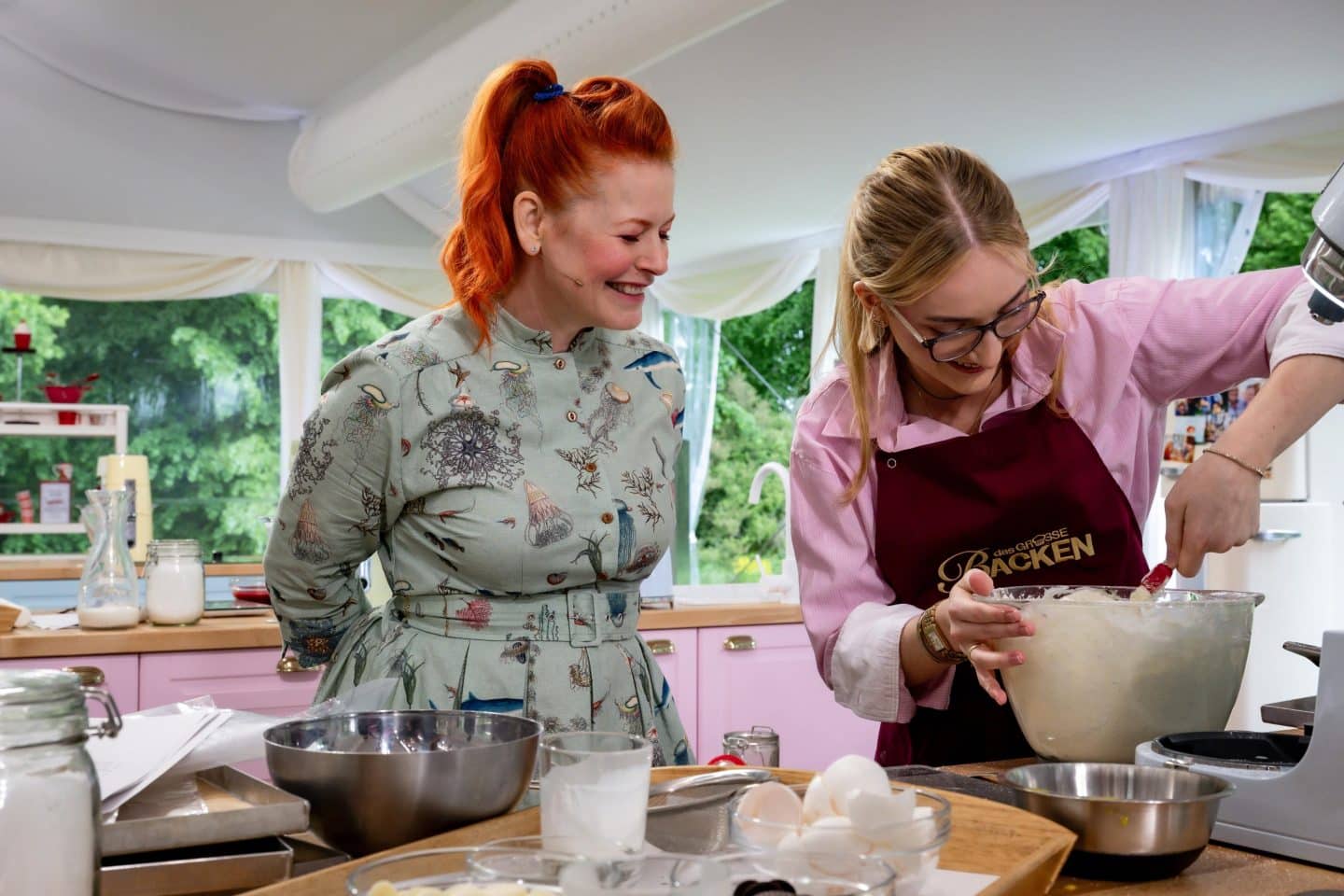 Enie van de Meiklokjes schaut lächelnd zu, wie Kandidatin Charlotte bei „Das große Backen“ Teig in einer Schüssel rührt.
