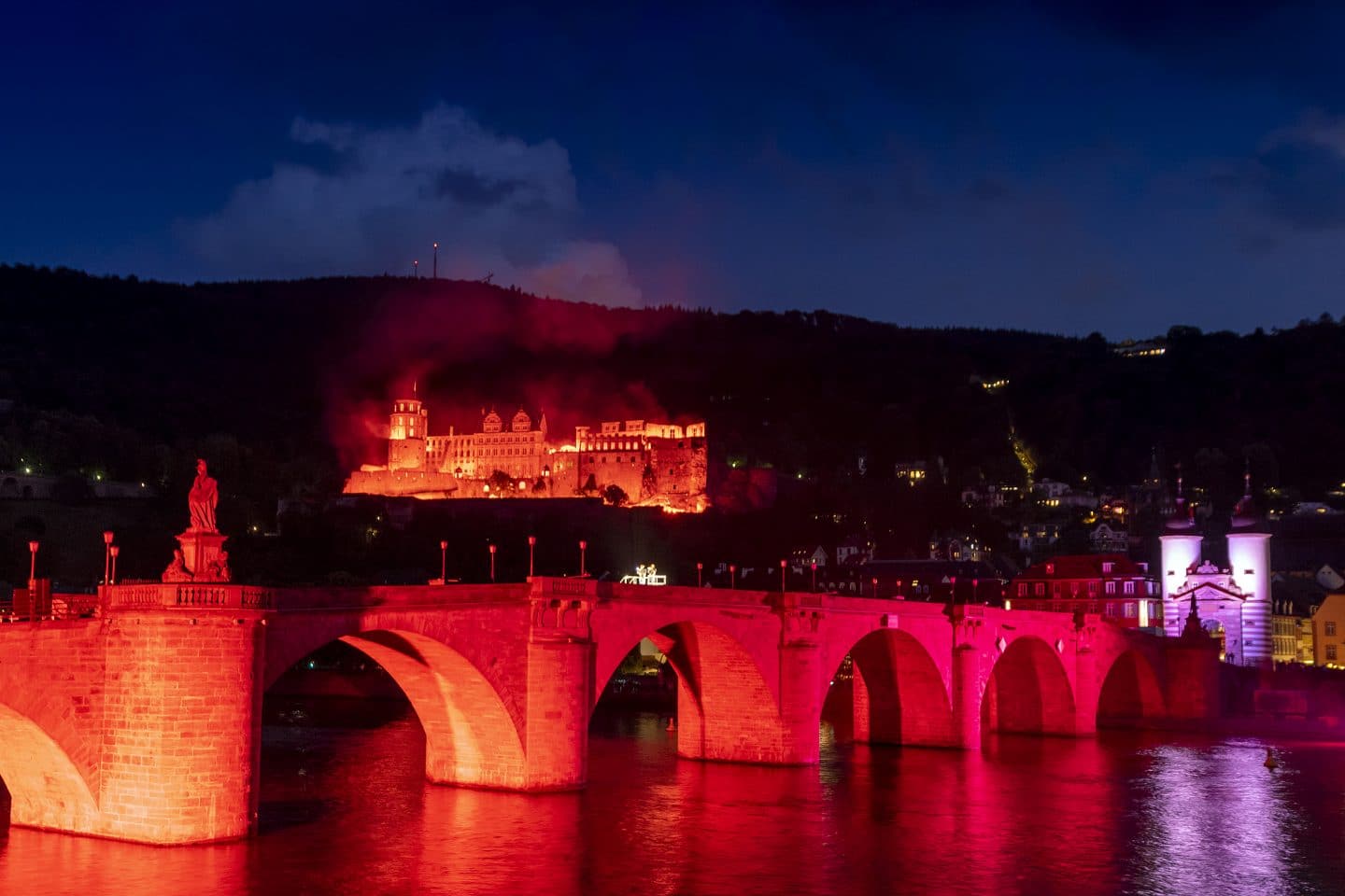 Das Heidelberger Schloss in rotem Licht bei Nacht, mit der hell erleuchteten Alten Brücke im Vordergrund.