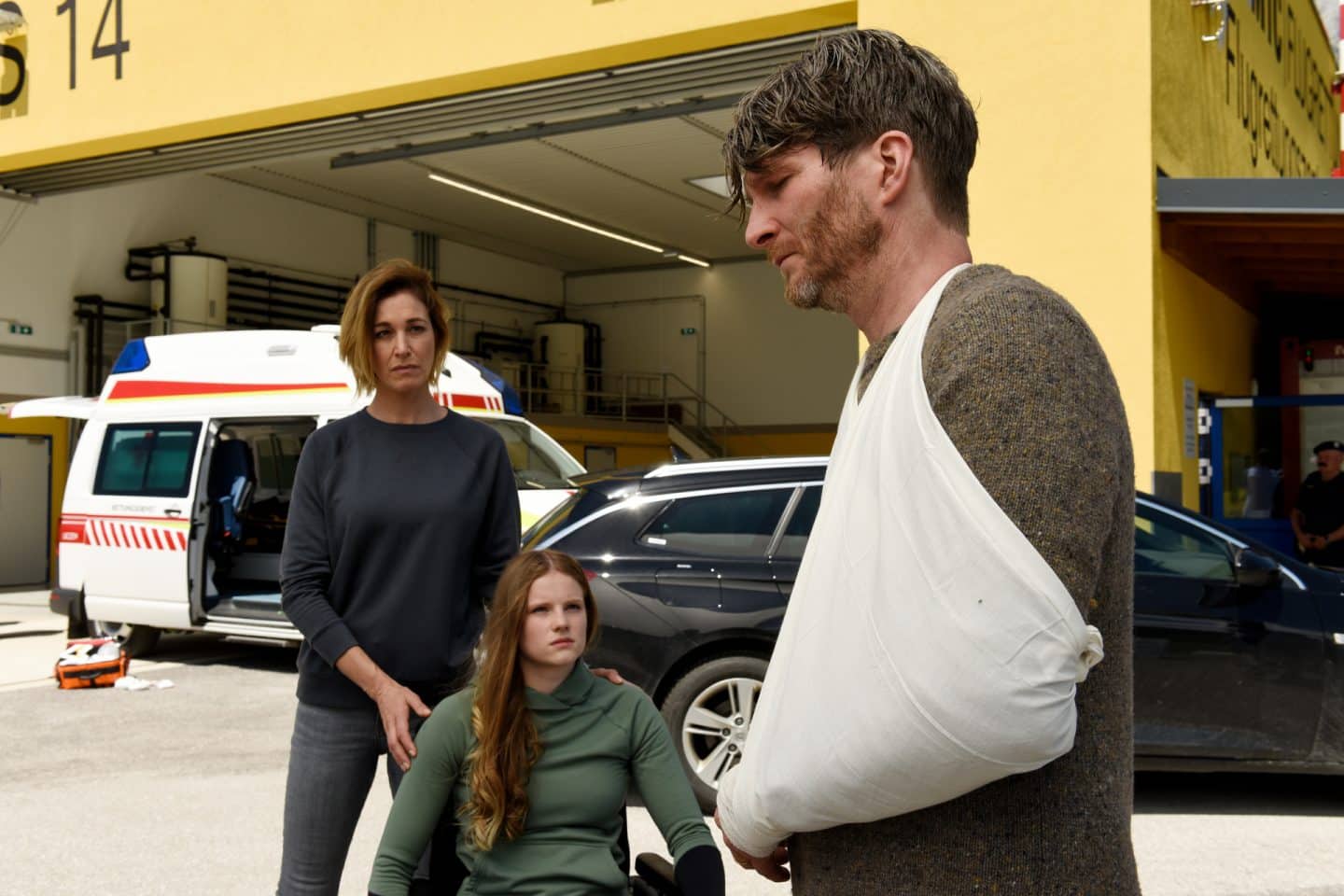 "Die Bergretter - Um jeden Preis": Julia Haas (Julia Stinshoff) steht hinter der im Rollstuhl sitzenden Pia (Emilia Warenski). Beide blicken fragend zu Georg (Leopold Hornung). Dieser hat den Arm in einer Schlinge gelegt und blickt traurig zu Boden. Im Hintergrund steht ein Krankenwagen.
