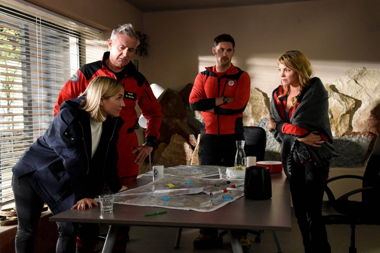 "Die Bergretter - Um jeden Preis": In der Einsatzzentrale stehen Jessi (Maxi Warwel), Michi (Robert Lohr), Simon (Ferdinand Seebacher) und Katharina (Luise Bähr) um einen Tisch herum. Auf diesem liegt eine ausgebreitete Landkarte. Alle sehen sich besorgt an. Katharina hat eine Decke um sich gelegt.

