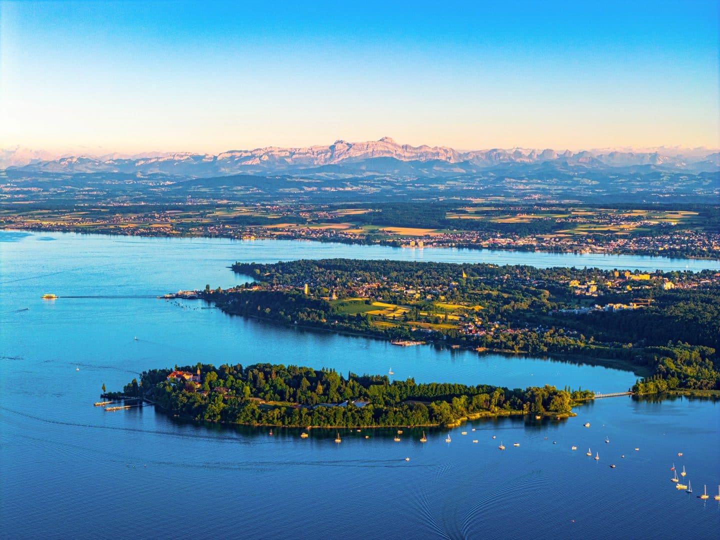 Luftaufnahme vom Bodensee mit der Insel Mainau und Alpenpanorama im Hintergrund.