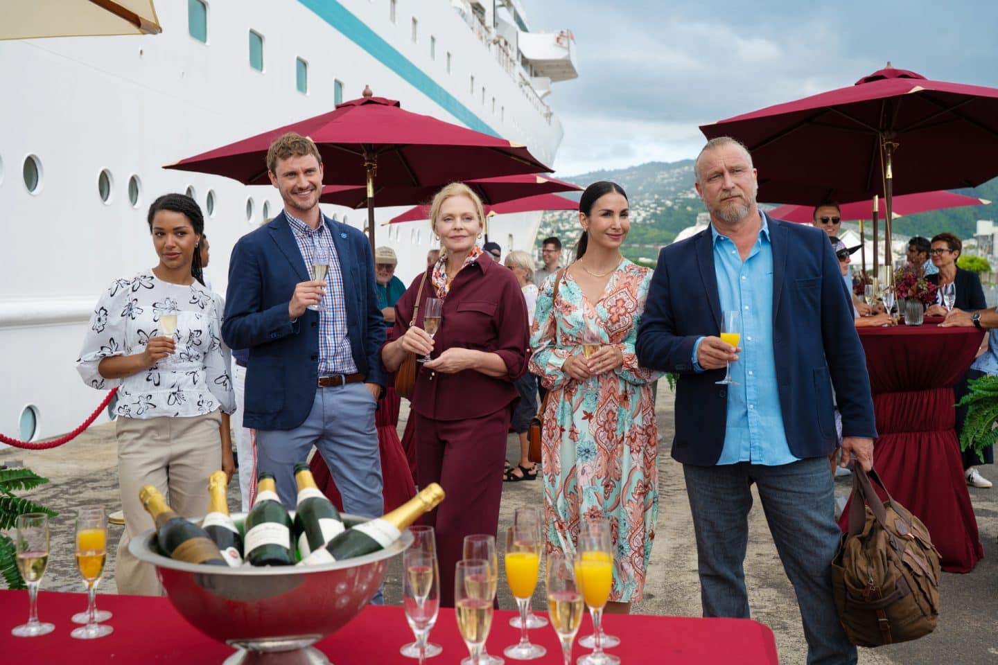 "Das Traumschiff: Auckland": Im Hafen: Esther Sarchow (Susanna Simon), Britta Weigert (Sila Sahin), Victoria Schneider (Luna Baptiste), Erik Milewski (Matthias Komm) und Marco Zurheide (Frédéric Brossier) stehen nebeneinander vor dem Schiff. Sie halten gefüllte Sektgläser in den Händen und wirken glücklich. Vor ihnen steht ein Tisch mit Sektflaschen und -gläsern. Im Hintergrund stehen weitere Passagiere (Komparsen) unter roten Sonnenschirmen an Stehtischen.
