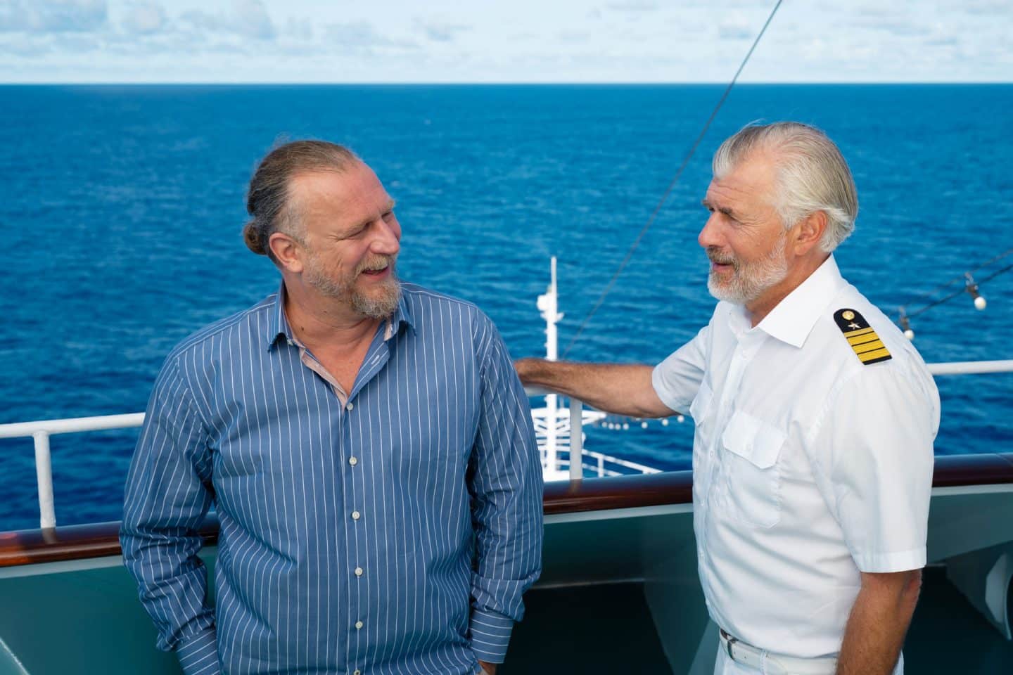 "Das Traumschiff: Auckland": An Deck des Schiffs: Am Geländer steht Erik Milewski (Matthias Komm) und blickt lachend zu Staff-Kapitän Martin Grimm (Daniel Morgenroth). Auch er ist gut gelaunt. Über dem offenen Meer sind einige Wolken am Himmel zu sehen.
