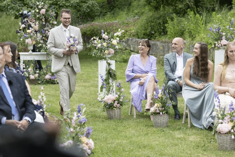 Ein Bräutigam im beigen Anzug mit Blumenstrauß läuft bei einer sommerlichen Outdoor-Hochzeit den Mittelgang entlang. Gäste sitzen auf beiden Seiten und lächeln ihm freudig entgegen.